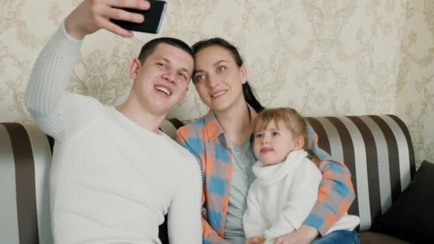A família caucasiana jovem feliz com a filha fotografa-se usando o smartphone em casa. Férias em família juntos foto de memória. Mãe, pai, filha de blogueiros se comunicam usando telefone moderno — Vídeo de Stock