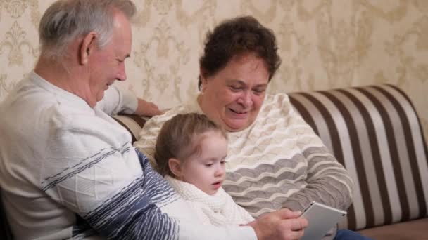 Lycklig familj, moderna morföräldrar leker med ett barn, en flicka som sitter med en surfplatta online hemma. Barn, flicka visar något i tabletten för morföräldrar. Äldre par med barnbarn och pryl — Stockvideo