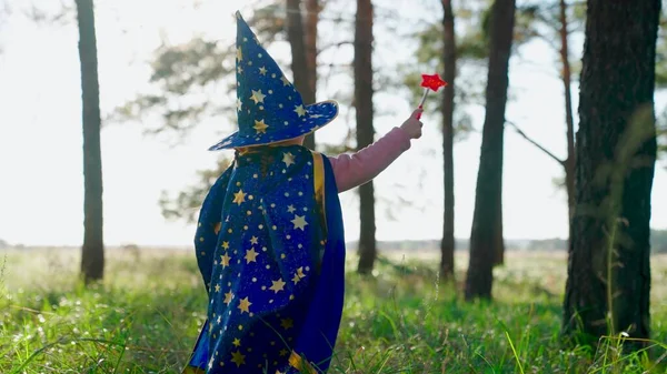 Kind fantaseert in mantel van goochelaar in het bos. Een meisje, kind speelt in een tovenaarskostuum, steekt haar hand op, zwaait met toverstaf in het park. Halloween. Gelukkige jeugd, familie. Kind spelen goochelaar buiten Stockfoto