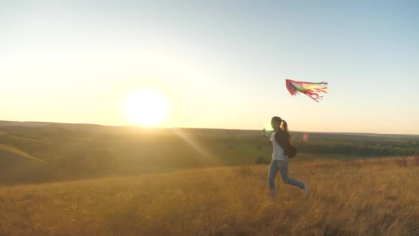 Szczęśliwa dziewczyna z latawcem w rękach przechodzi przez pole w promieniach zachodzącego słońca. Beztroskie dziecko marzy o wolności, ucieczce. Nastolatek bawi się zabawkami na świeżym powietrzu w parku. Koncepcja dziecięcych marzeń — Wideo stockowe
