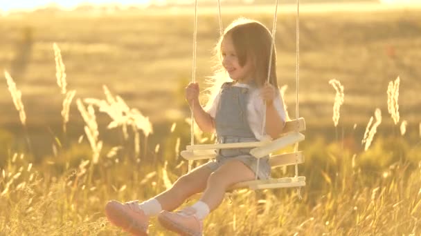 Feliz niño, niña, balanceándose en un columpio bajo un árbol en el sol en el parque. Feliz infancia. Una niña vuela en un columpio en un patio de recreo. La niña, la hija se ríe, se regocija y agita los brazos en vuelo — Vídeos de Stock