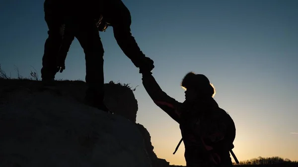 旅行者はお互いに崖に登るのを助ける。登山家のシルエットは、山の頂上に登るお互いに手を差し伸べます。ビジネスマンのチームワーク。ビジネスマンのチームが勝利に行く. — ストック写真