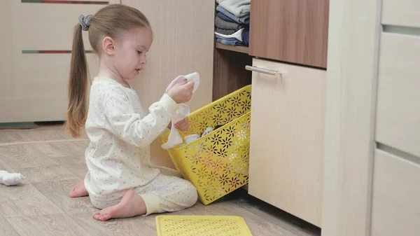Маленькая девочка в пижаме открывает шкаф и ищет свою одежду. Счастливая маленькая девочка играет в детской комнате с одеждой. Парень выбрасывает свои вещи из коробки. Дочь играет с одеждой дома — стоковое фото