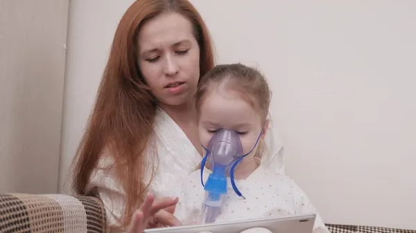 Mamá trata a su hija con la inhalación juntos ven dibujos animados en la tableta. Familia tratando a un niño en casa por resfriados. El niño sufre de tos. El niño está recibiendo terapia respiratoria con nebulizador. — Foto de Stock