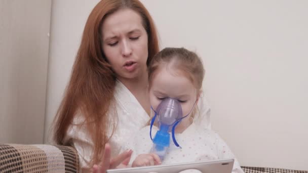 Mamá trata a su hija con la inhalación juntos ven dibujos animados en la tableta. Familia tratando a un niño en casa por resfriados. El niño sufre de tos. El niño está recibiendo terapia respiratoria con nebulizador. — Vídeos de Stock