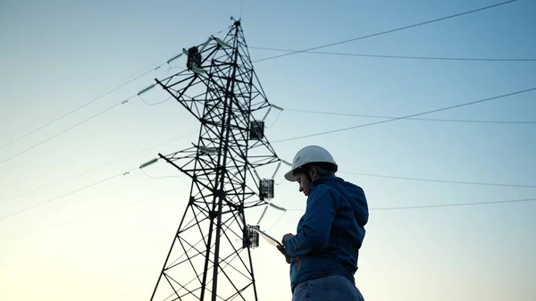 Ingénieur électricien femme ingénieur en casque de protection travaille à l'extérieur Lignes électriques haute tension au coucher du soleil. Distribution, fourniture d'électricité. Énergie électrique respectueuse de l'environnement. Électricien Photo De Stock