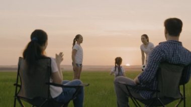 Mutlu aile, anne, baba, tatildeki çocuklar gün batımında parkta top oynuyorlar. Anne ve baba tarlada turist sandalyelerinde oturuyorlar, çocuklar güneşle, çocuklukla, tatil seyahatleriyle oynuyorlar.