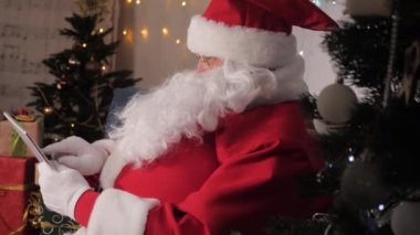 Santaclaus dijital tabletle çalışıyor. Modern aletler Noel Baba 'nın elinde. Noel zamanı. Yeni yıl aile tatili. Noel Baba tablet bilgisayar kullanır. Listesini çocuklar için hediyelerle kontrol eder. Noel Baba, teknoloji.