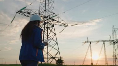 Kadın elektrik tedarik şirketi çalışanı dışarıda çalışıyor, günbatımında yüksek voltajlı elektrik hatlarına hizmet veriyor. Beyaz miğferli mühendis kadın güç mühendisi güç hattını bilgisayar tableti kullanarak kontrol ediyor.