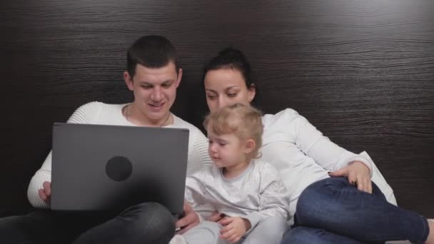 Córka ogląda kreskówkę z mamą i tatą, używa laptopa, siedzi na łóżku. Szczęśliwa rodzina z małym dzieckiem uczy się w domu na komputerze online, słodkie dziecko bawi się z rodzicami. — Wideo stockowe