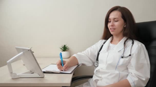 女医生穿着白衣、听诊器工作，在医院办公室使用电脑平板为病人提供远程咨询。家庭医生在网上谈话。远程医疗。医生工作 — 图库视频影像