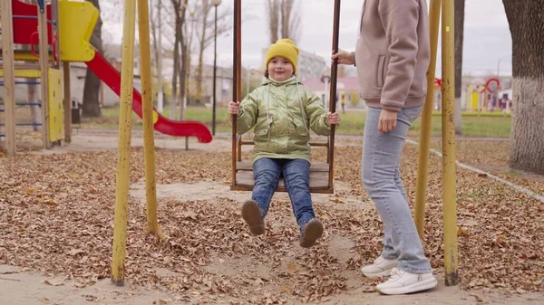 Mutlu genç anne ve küçük kızı sonbaharda parkta salıncakta sallanıyor. Güzel küçük kız salıncakta oturuyor. Anne ve kızı oyun alanında birlikte oynuyorlar. Mutlu aile kavramı. Aile dışarıda yürür. — Stok fotoğraf