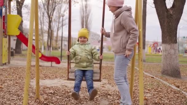 Šťastná mladá matka a dcera houpačka na houpačce v parku na podzim. Krásná holčička sedí na houpačce. Máma a dcera si spolu hrají na hřišti. Šťastný rodinný koncept. Rodinné procházky venku — Stock video