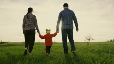 Mutlu aile yeşil çimlerde parkta yürüyor, baharda gün batımında el ele tutuşuyorlar. Mutlu çocukluk, aile eğitimi anne, baba ve kız sahada oynuyorlar, çocuk ebeveynleri elleriyle tutuyor. Takım çalışması..