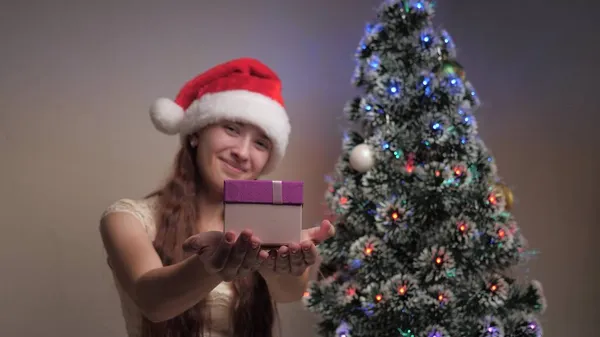 메리 크리스마스. 산타클로스 모자를 쓴 한 소녀가 크리스마스 트리 옆에 선물을 들고 있습니다. 십 대들은 선물 상자를 들고 카메라를 들고 기뻐 하며 미소를 짓는다. 가족의 아이들의 휴일 과 축하, 겨울 방학. — 스톡 사진