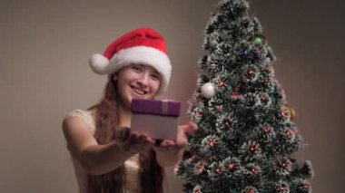 Mutlu noeller. Noel Baba şapkalı bir kız Noel ağacının yanında hediye tutuyor. Ergen, kameraya hediye kutusunu uzatır, sevinir ve gülümser. Aile çocukları tatil, kutlama, kış tatili.