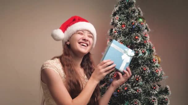 Szczęśliwa dziewczyna trzyma pudełko prezentów świątecznych w rękach, raduje się i uśmiecha. Dziewczyna w kapeluszu Świętego Mikołaja trzyma prezent obok choinki. Rodzinne zimowe wakacje dla dzieci. Wesołych Świąt. — Wideo stockowe