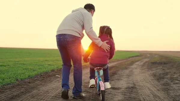 Happy family, dad teaches his daughter, child to ride a bike in the park at sunset. Father teaches a little kid girl to ride a childs bike on the road, in the fall, in spring. Happy family, childhood