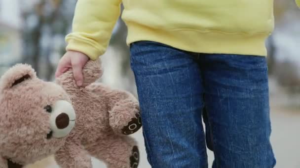Een klein kind met een teddybeer in zijn hand close-up tijdens een wandeling in het herfstpark. Kind met een teddybeer op de speelplaats. Eenzaam kind met teddybeer buiten. Teddy beer in handen van zoon, dochter — Stockvideo