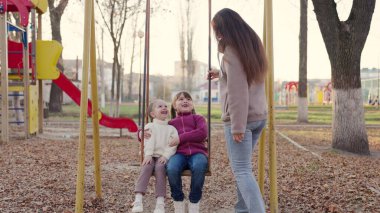 Mutlu anne ve küçük kızları sonbaharda parkta salıncakta sallanıyor. Güzel küçük kızlar salıncakta oturup gülerler. Anne, çocuklar parkta birlikte oynuyorlar. Mutlu aile kavramı