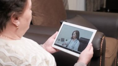 Yaşlı bir kadın, sağlık hizmeti sağlayıcısıyla konuşmak için dijital tablet kullanıyor. Tıbbi video konferans uygulaması. Profesyonel bir kadın doktor bir hastayı internetten görüntülü konuşma ile tedavi ediyor.