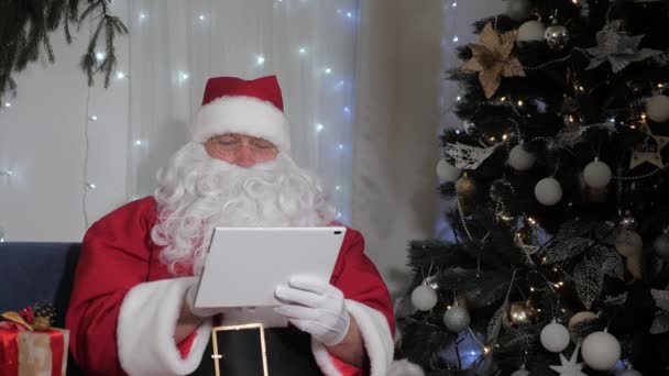 サンタクロースはデジタルタブレットで動作します。サンタの手にある現代のガジェット。クリスマスだ。新年の家族の休日。サンタクロースはタブレットコンピュータを使用し、子供への贈り物で彼のリストをチェックします。サンタ、テクノロジー — ストック動画