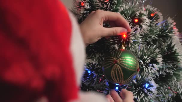 Child girl in red Santa Claus hat decorates the Christmas tree. Close-up. Kid hangs a green toy ball on the tree. Happy family, childhood, Christmas. Teenager decorates a Christmas tree, New Year. — Stock Video