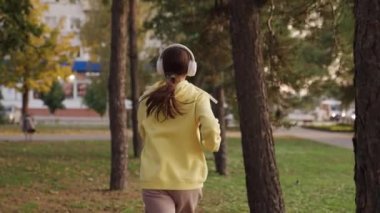Kadın, koşu, spor, güç. Şehir parkındaki ağaçların arasında koşan sporcu kız. Genç bir kadın, aktif bir atlet kulaklık takıyor, müzik dinliyor, koşuyor, parkta antrenman yapıyor. Koş ve kilo ver.