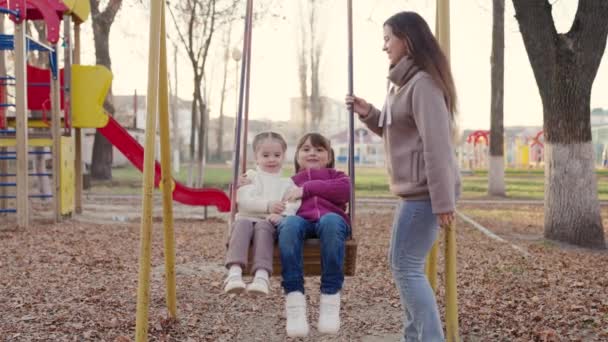Boldog fiatal anya és kislányai hintáznak a parkban ősszel. Gyönyörű kislányok ülnek a hintán és nevetnek. Anya, a gyerekek együtt játszanak a játszótéren. Boldog családi koncepció — Stock videók