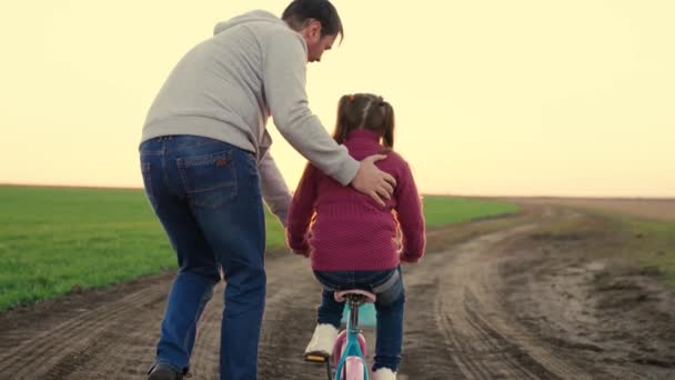 Счастливая семья, папа учит свою дочь, ребенка ездить на велосипеде в парке на закате. Отец учит маленькую девочку кататься на детском велосипеде по дороге, осенью, весной. Счастливая семья, детство — стоковое видео