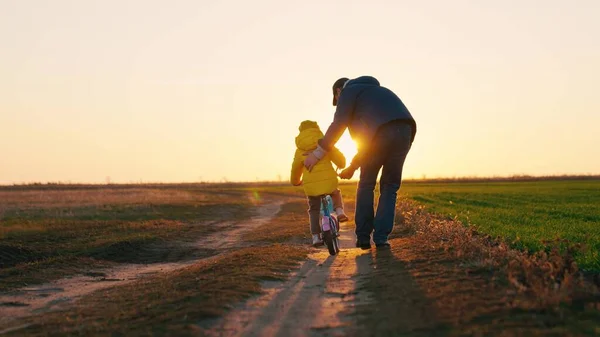 Father teaches a little kid girl to ride a childs bike on the road, in the fall, in spring. Happy family, childhood. Happy family, dad teaches his daughter, child to ride a bike in the park at sunset