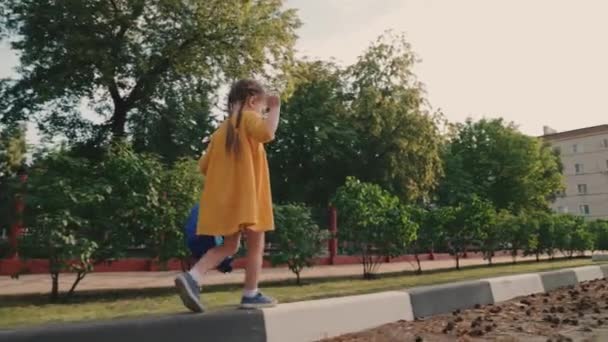 Dziecko, mała dziewczynka z plecakiem wraca do domu ze szkoły i bawi się na ulicy. Sny z dzieciństwa. Dzieciak uczy się zachować równowagę. Mała dziewczynka spaceruje po letnim parku. Rodzina, wesołe dzieciństwo — Wideo stockowe