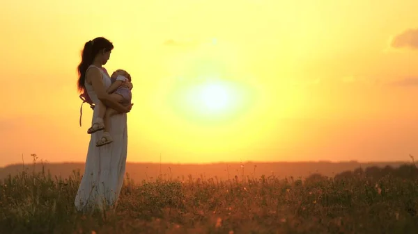 Mamãe abraça sua filha, o bebê quer dormir, eles admiram o belo pôr do sol no campo. Família feliz, criança e pais. Mãe e filho estão andando no parque, no verão ao sol. — Fotografia de Stock