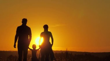 Mutlu aile ve çocuk yazın gün batımında parkta yürüyorlar. Takım çalışması. Ebeveynler ve kızları güneşin altında el ele yürüyorlar. Mutlu bir ailenin silueti açık havada birlikte seyahat ediyor.
