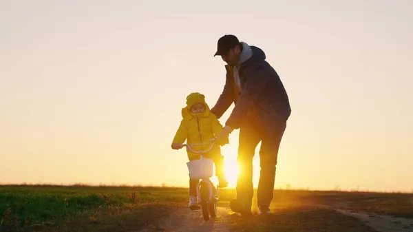 O pai ensina a criança a andar de bicicleta no parque ao pôr-do-sol, família feliz. Pai ensina uma menina, filha a andar de bicicleta infantil na estrada no outono, primavera. Infância feliz, sonhos de infância — Fotografia de Stock