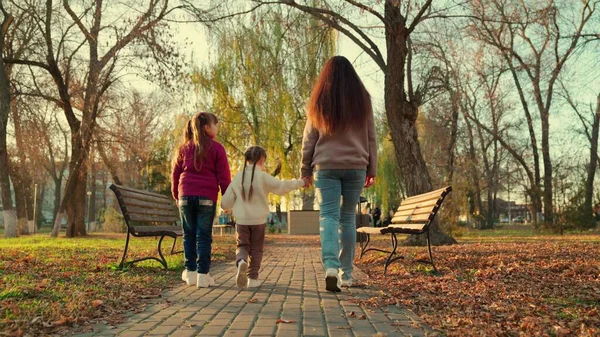 妈妈带着两个孩子在日落时散步公园，快乐的家庭，孩子牵着慈母的手，女孩子们和她们的父母一起散步秋天的城市公园，黄叶是季节的时候，快乐的母爱 — 图库照片