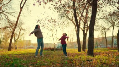 Annesiyle birlikte küçük bir çocuk sonbahar parkında günbatımında sarı yapraklar fırlatıyor, mutlu bir aile, annesiyle çocuk sonbaharda altın yaprak döküyor, eğlenceli bir oyun fantezisi kuruyor, gün ışığında eğleniyor.