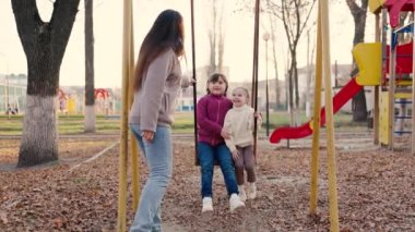 Anne, sonbahar parkında sallanan küçük kızları sallıyor mutlu bir aile, yürüyüş parkında çocuklar gülüyor, çocuk eğleniyor, gülümsüyor, uçuyor, dinleniyor sağlıklı dinleniyor, eğlenceli yürüyüşler yapıyor.