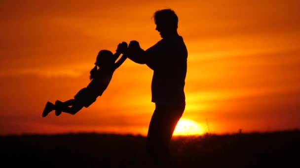 Szczęśliwa rodzina, tata i córka bawią się razem na polu o zachodzie słońca. Tata okrąża swoją ukochaną córkę dłońmi w promieniach słońca. Ojciec córka spaceruje razem w parku w lecie — Wideo stockowe
