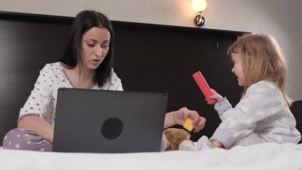 Mamma lavora con un computer portatile e gioca con i giocattoli con la sua figlioletta. La mamma multitasking lavora da remoto a casa sul computer e si prende cura del bambino. Problemi legati al lavoro a domicilio — Video Stock