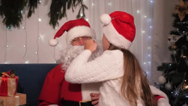 Papai Noel está brincando com uma menina sentada em seu colo. Miúda, o miúdo pede um desejo ao Santaclaus. Véspera de Natal, conceito de férias de crianças de família. Festa de Natal em casa. Feliz feriado de inverno — Vídeo de Stock