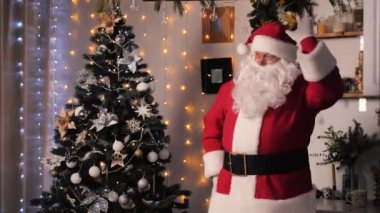 Noel arifesinde, Noel Baba süslenmiş bir dairede topları ve ışıkları olan bir Noel ağacının yanında dans ediyor. Aile kavramı, çocuk tatili. Neşeli Noel Baba evde yeni yılı kutluyor..