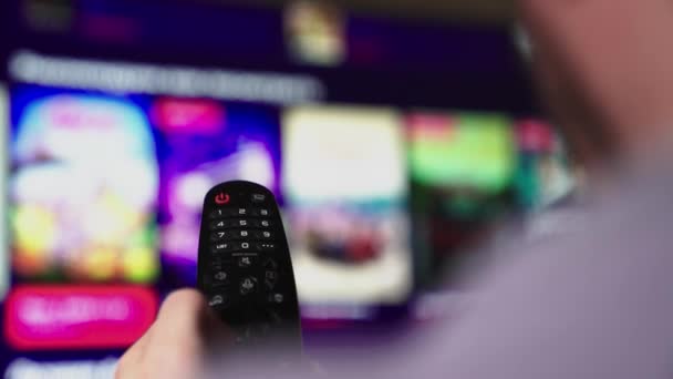 Een man kijkt smart TV en gebruikt zwarte afstandsbediening. Wazige tv scrollt pagina 's. Mans hand selecteert internet tv-kanalen met afstandsbediening, close-up. Persoon bestuurt TV met behulp van een moderne afstandsbediening. — Stockvideo