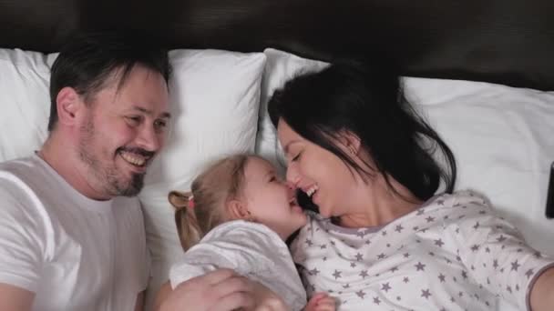 Familia joven feliz con su hija son fotografiados en un teléfono inteligente en la cama. Madre, padre, hijo, la niña, jueguen juntos en el sofá de casa. Concepto de familia joven moderna — Vídeos de Stock