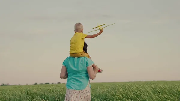 Ένας γιος στους ώμους μιας μητέρας, περπατούν μαζί σε ένα χωράφι με πράσινο σιτάρι. Το αγόρι παίζει με τη μητέρα του, κρατάει ένα παιχνίδι αεροπλάνο στο χέρι του. Η παιδική ηλικία ονειρεύεται να γίνει πιλότος. Οικογένεια — Φωτογραφία Αρχείου