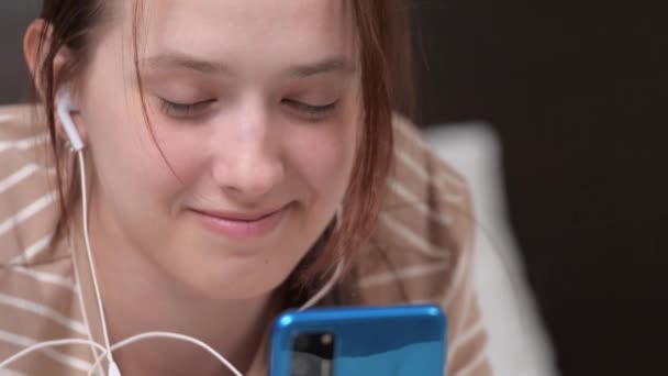 Uma jovem com fones de ouvido se comunica usando seu smartphone na Internet, ouve música, navega em redes sociais, joga jogos, envia mensagens de texto, sentada no sofá em casa. Close-up — Vídeo de Stock
