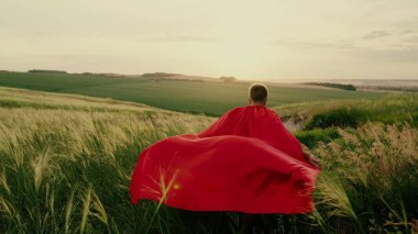 Mutlu bir çocuk, süper kahraman bir çocuk, kırmızı bir pelerinle yeşil alanda koşuyor, pelerin rüzgarda dalgalanıyor. Çocuk oyunları, rüyalar. Ağır çekim. Gençler süper kahraman olmayı hayal eder..