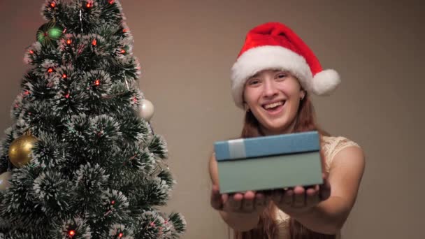 Wesołych Świąt. Dziewczyna w kapeluszu Świętego Mikołaja obok choinki trzyma prezent. Szczęśliwa dziewczyna trzyma pudełko prezentów świątecznych w rękach, raduje się i uśmiecha. Rodzinne zimowe wakacje dla dzieci. — Wideo stockowe