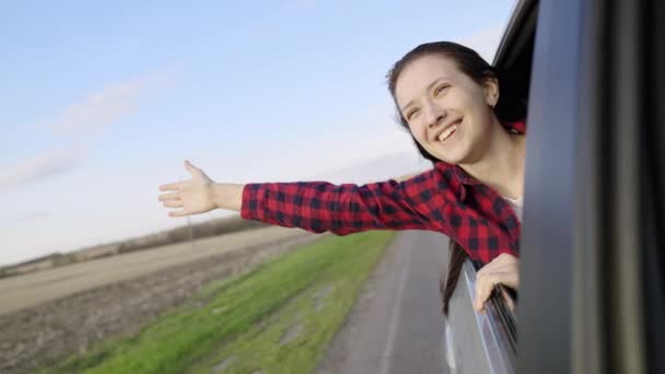 長い髪を持つ若い女性は、笑顔、車の後部座席に座って、車の窓から幸せと夢をキャッチします。無料の女の子は車の窓から手で風をキャッチ車で移動します。十代の旅 — ストック動画
