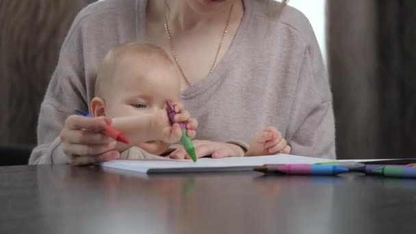 Mãe, o bebé brinca e desenha em casa no quarto. Jovem mãe ensina filho bebê bonito para desenhar com marcadores coloridos na folha de papel. Família adere à educação infantil sentado à mesa no quarto em casa. — Vídeo de Stock